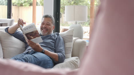 Feliz-Hombre-Birracial-Mayor-Relajándose-En-El-Sofá-Leyendo-Un-Libro-En-La-Sala-De-Estar,-Copiando-Espacio,-Cámara-Lenta