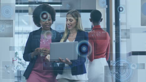 professional women sharing laptop in office corridor, displaying floating tech interface graphics