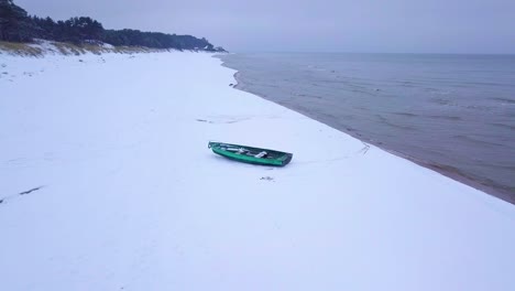 Vista-Aérea-De-La-Costa-Del-Mar-Báltico-En-Un-Día-De-Invierno-Nublado-Con-Un-Barco-De-Pescadores-Costero-Verde,-Playa-Con-Arena-Blanca-Cubierta-De-Nieve,-Erosión-Costera,-Tiro-De-Drones-En-órbita-De-Gran-Angular