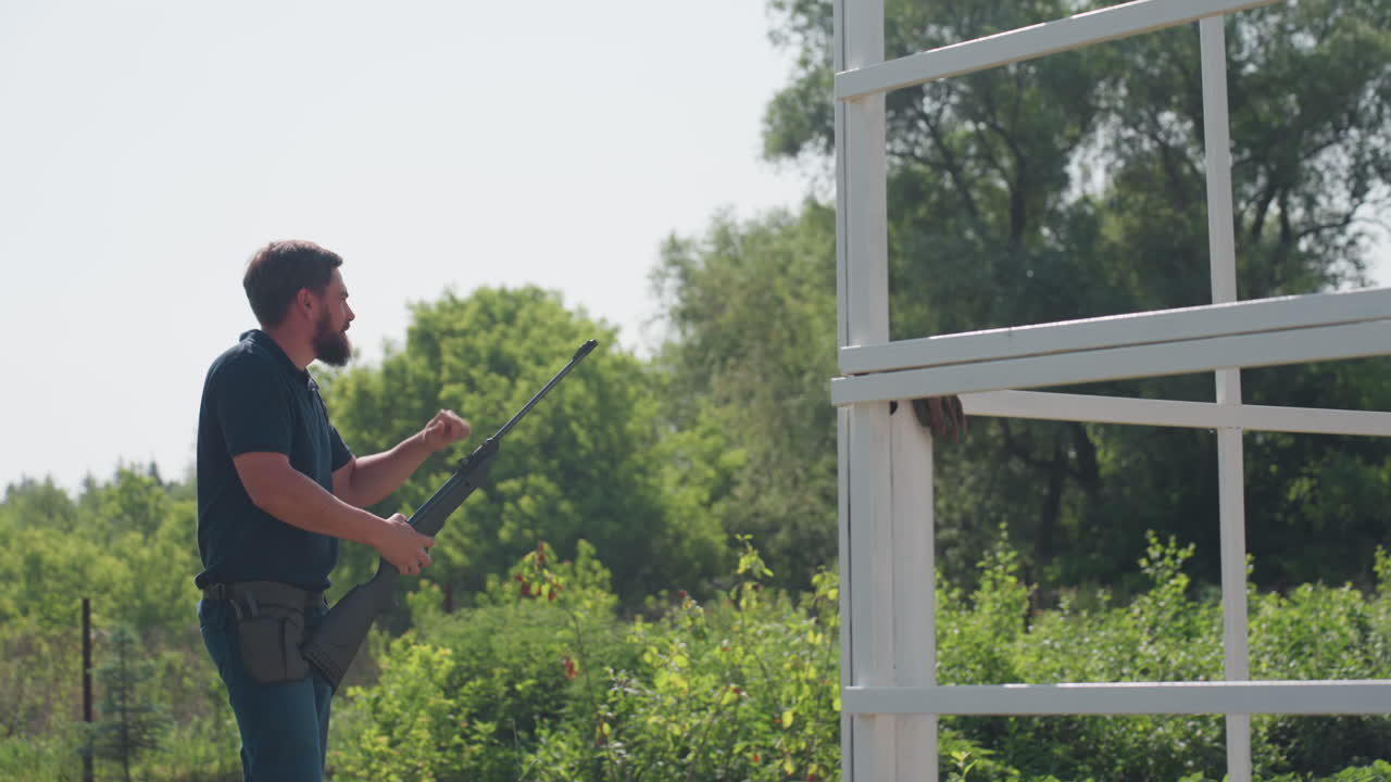 close up drum in foreground, side view of man aiming rifle toward distant target near farm frame, rural greenery background, protective action to guard crops from pests