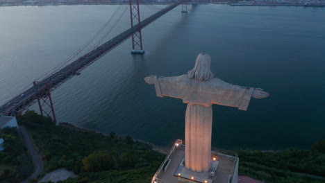 Close-up-aerial-orbit-of-large-white-monument-of-Santuario-de-Cristo-Rei-and-red-Ponte-25-de-Abril-bridge-off-the-coast-of-Lisbon,-Portugal