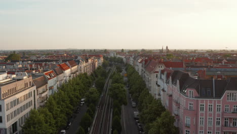 Forwards-fly-above-wide-street-with-railway-tracks-and-trees-in-city.-Morning-shot-of-urban-neighbourhood.-Berlin,-Germany