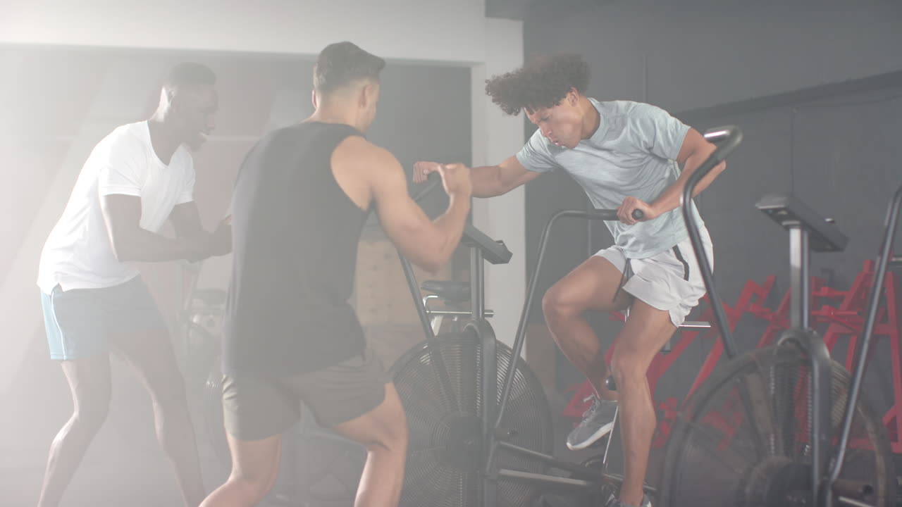 Three men exercising in gym, one using exercise bike while two others encouraging