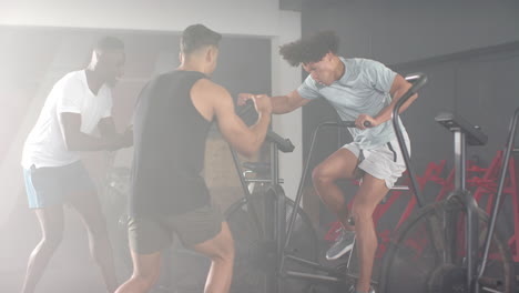 three men exercising in gym, one using exercise bike while two others encouraging