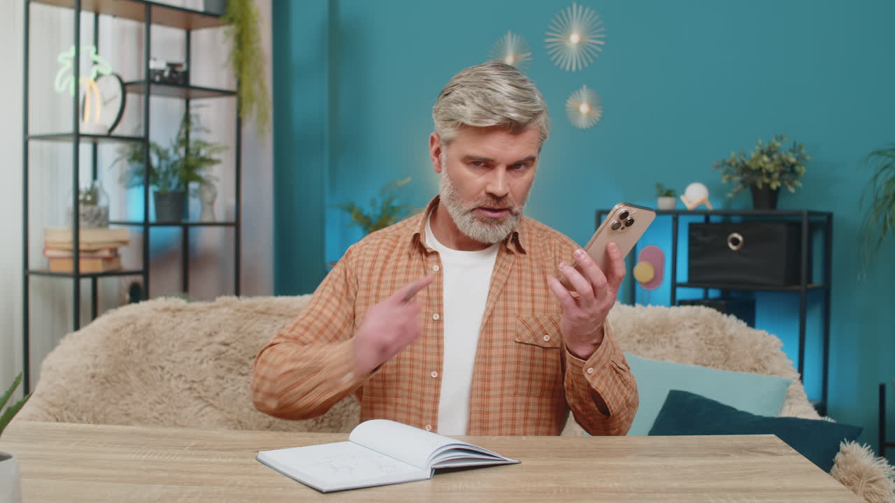 Uomo maturo al tavolo che parla al telefono in vivavoce, prende appunti sul quaderno, serio, a casa