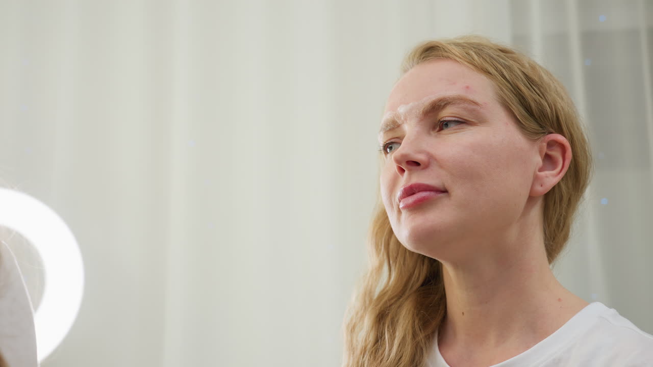 Lady with blonde hair tucked behind ear sits calmly as beautician gently cleans her eyebrow using cotton pad during beauty session under soft lighting in modern salon environment