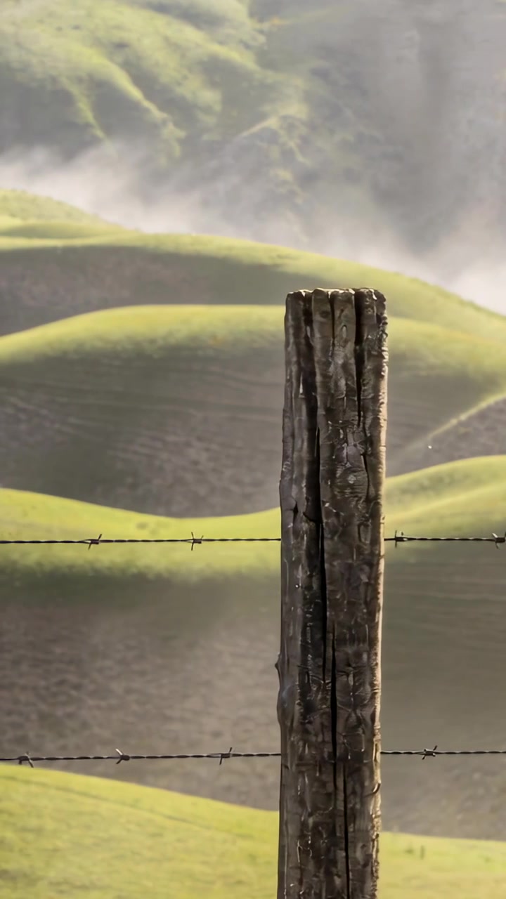 Video verticale: Telecamera panoramica che ricentra un palo consumato dalle intemperie tra colline nebbiose e filo spinato, spazio per il testo.