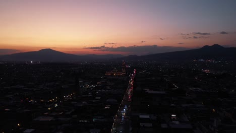 Drone-captures-Morelia,-Michoacán-at-dawn-with-car-lights-tracing-the-city’s-arteries-beneath-a-pastel-sunrise-sky