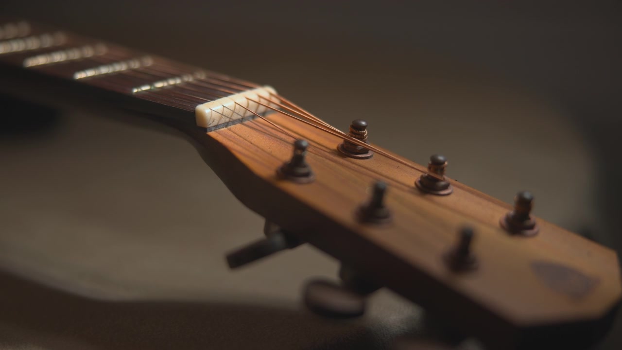 Close-up of an acoustic guitar headstock with tuning pegs on the right side, softly blurred against a dark background with warm low lighting and shallow depth