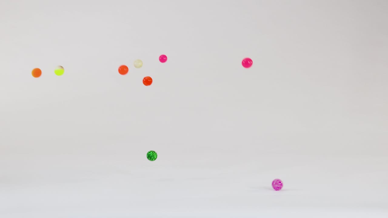 Bola karet multi-warna jatuh, memantul, dan tersebar di latar belakang studio putih bersih dalam gerakan lambat.
