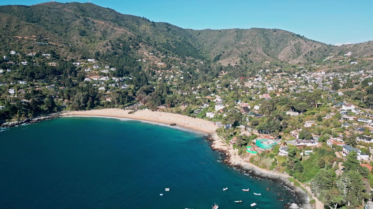 Amplia toma panorámica sobre una bahía de azul profundo con barcos anclados revelando una playa de arena en forma de media luna y un pueblo exclusivo anidado entre colinas costeras boscosas. Zapallar, Chile