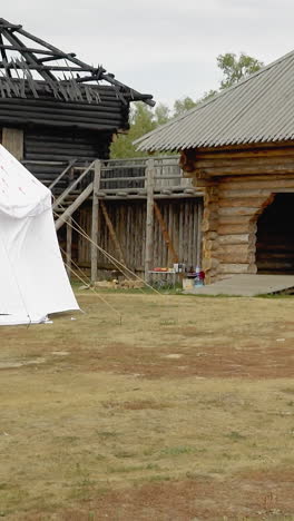 courtyard of medieval settlement with wooden gates and white tents. log building with burnt roof in ancient village under cloudy sky. place of historical memory