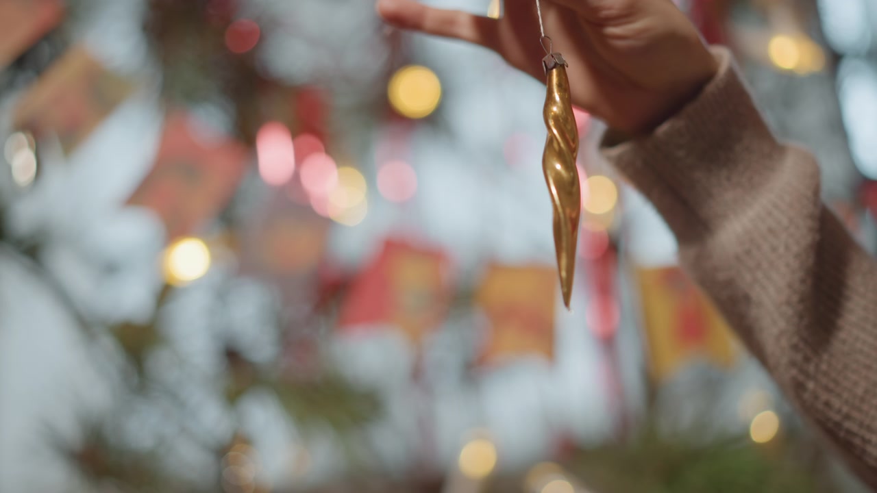 Ornamento a ghiacciolo dorato tenuto contro una manica di maglione morbida con effetto bokeh, ghirlanda colorata e lucine calde, posizionamento delicato su un ramo sempreverde, atmosfera accogliente e nostalgica, movimento cinematografico lento.