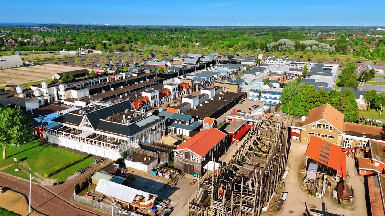 Flying closer to the berth with the boats and ship near it. Museum Batavialand in Lelystad, the Netherlands. Aerial view on the green city on sunny day.
