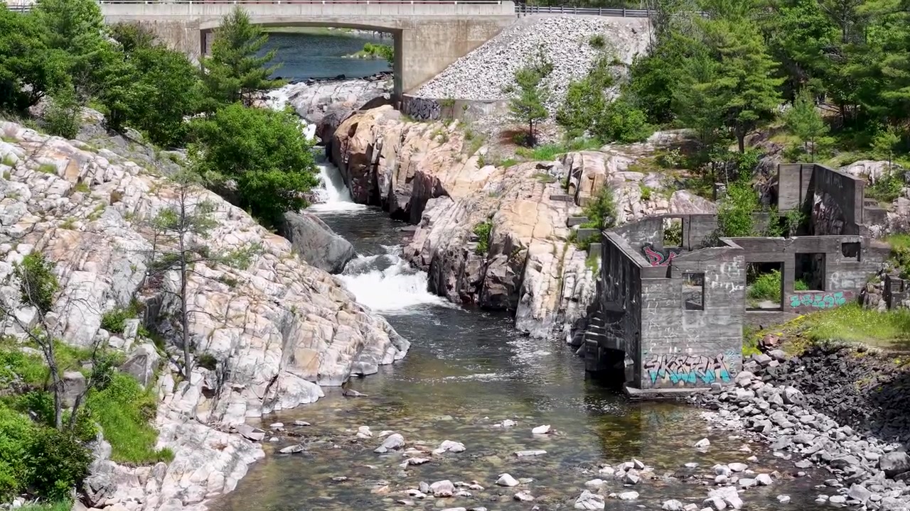Whitefish Şelaleleri yakınında, terk edilmiş, grafiti kaplı beton endüstriyel bir harabenin yanındaki granit bir kanyonda coşkulu şelalelere yaklaşan drone.
