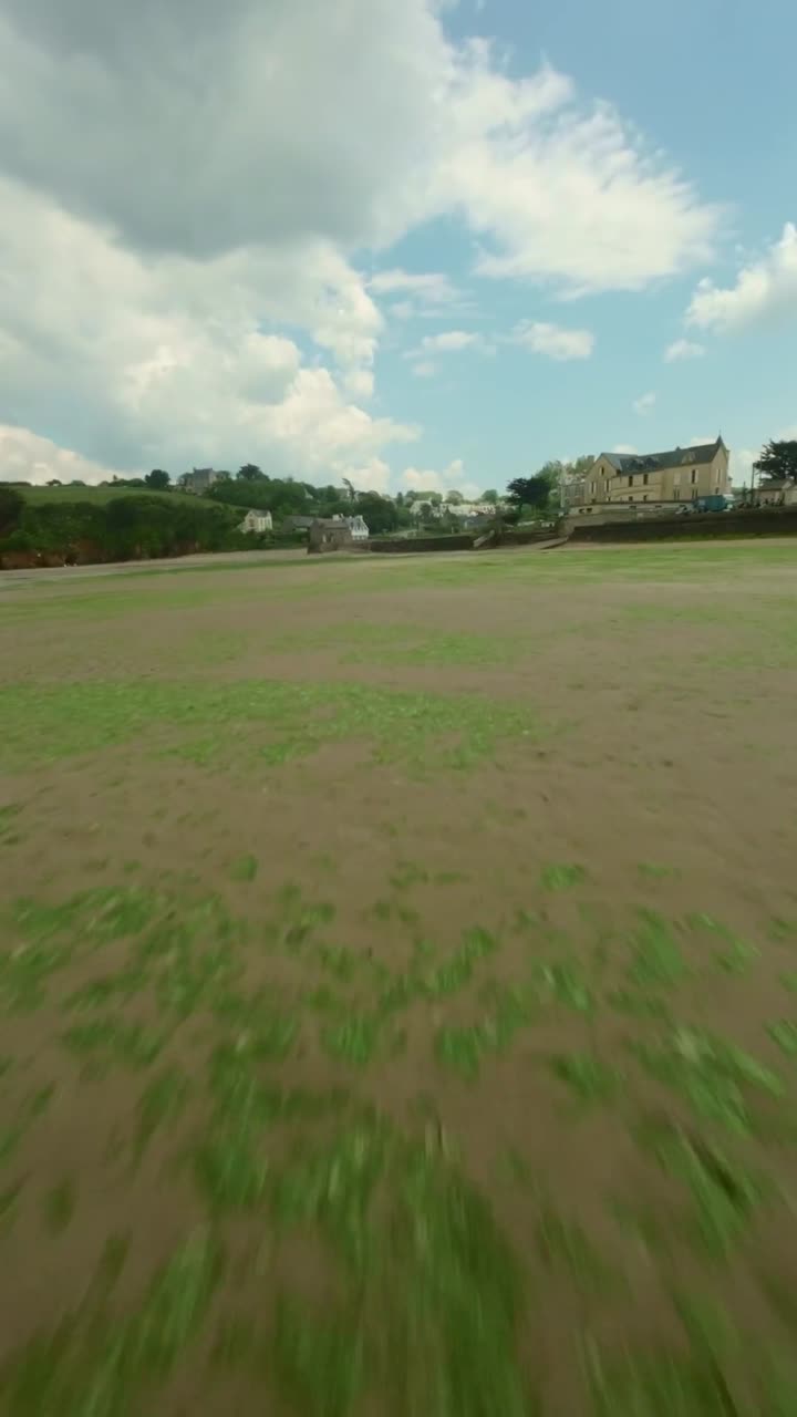 FPV drone races along sandy Brittany shoreline with soft waves and coastal cliffs under bright sky, tourists on beach