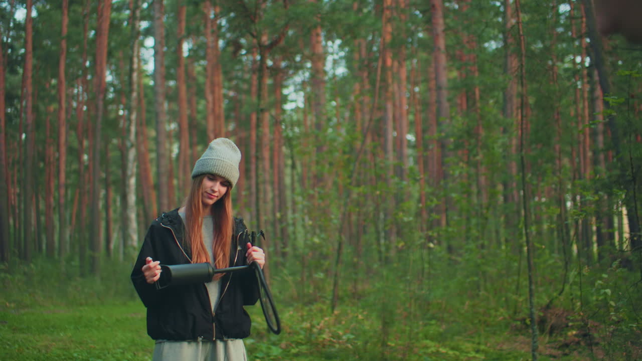 Close up of man spraying tent fabric in forest as woman beside him holds and plays with air pump during outdoor camping session surrounded by pine trees and natural greenery