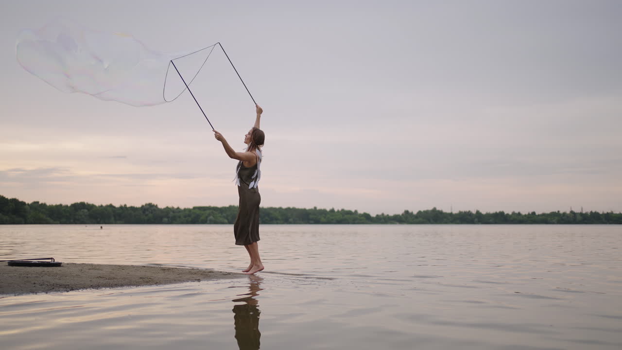 una joven artista muestra un espectáculo de burbujas de jabón haciendo estallar enormes burbujas de jabón en la orilla de un lago al atardecer. muestra un hermoso espectáculo de burbujas de jabón en cámara lenta