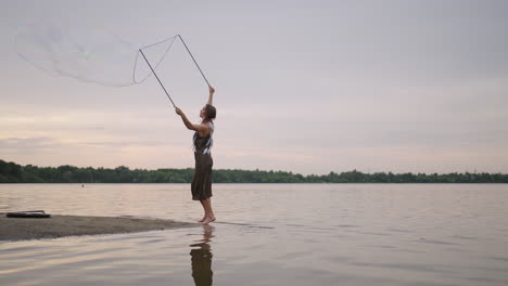 A-young-female-artist-shows-a-soap-bubble-show-blowing-up-huge-soap-bubbles-on-the-shore-of-a-lake-at-sunset.-Show-a-beautiful-show-of-soap-bubbles-in-slow-motion