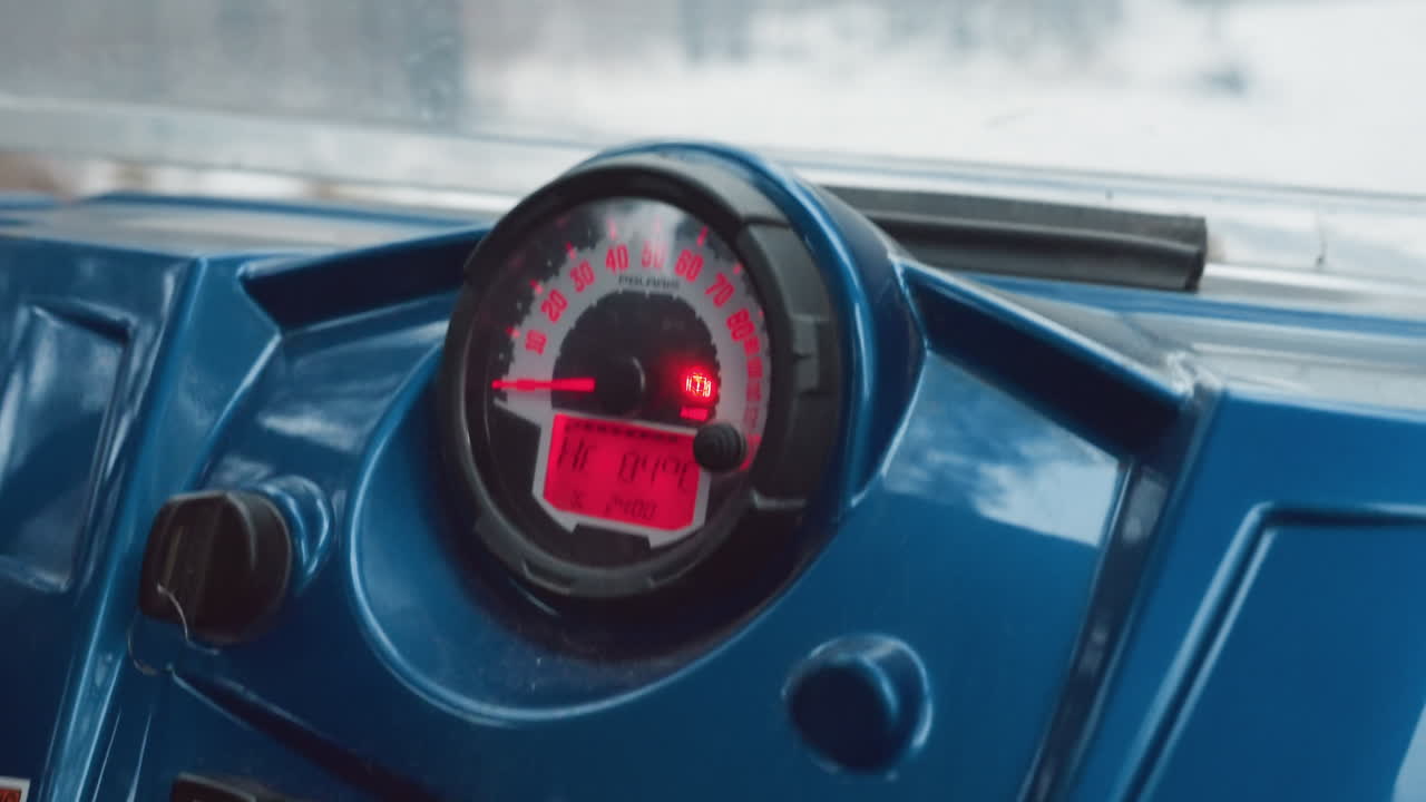 Atv dashboard speedometer glowing red pilot grips steering, throttle engaged, snowcovered trail visible through windshield, cold overcast light, instrument panel and digital meter, closeup of rpm