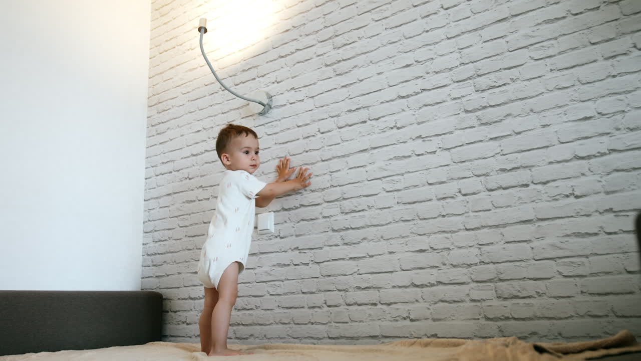 Lovely Caucasian toddler boy in white bodysuit stands on the bed. Little kid plays happily with lamp. Low angle view.
