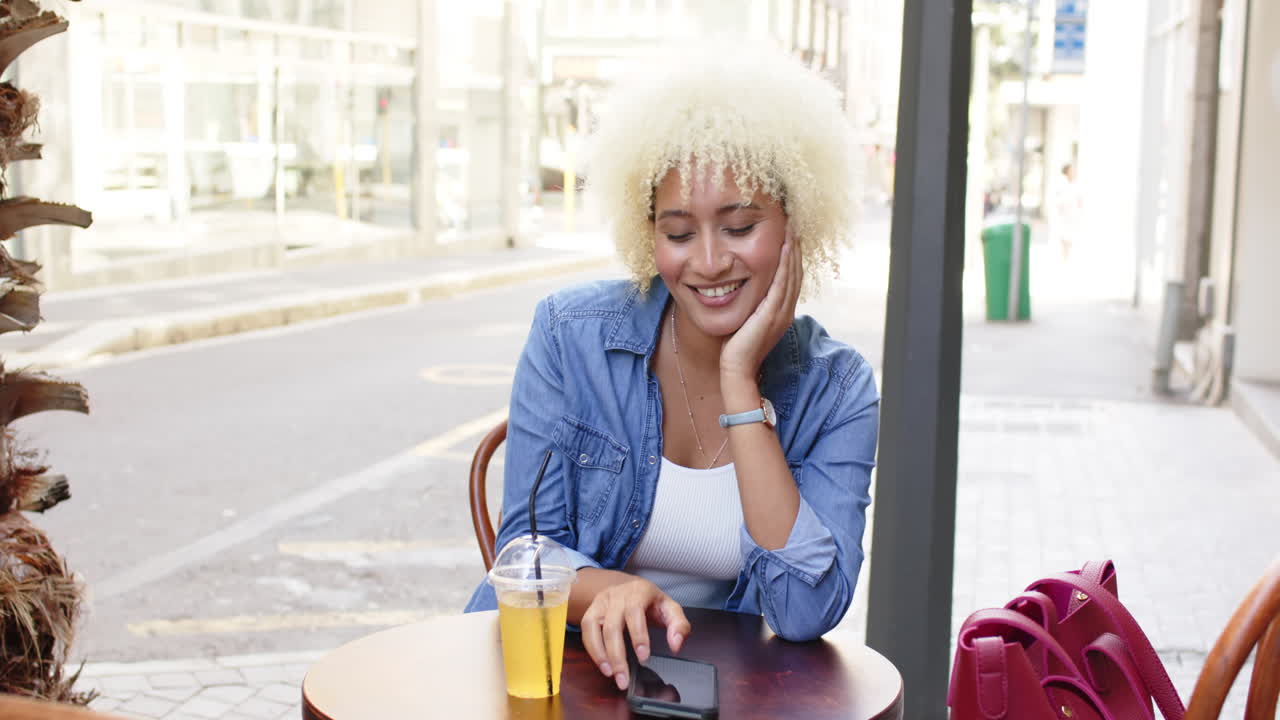 Young biracial woman with curly blonde hair enjoys a beverage at an outdoor cafe
