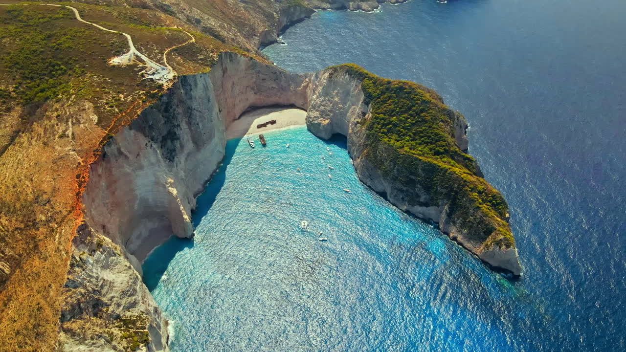 Aerial drone view of the Navagio beach on the Ionian Sea coast of Zakynthos, Greece. Rocky cliffs, moored and floating boats, resting people, blue water