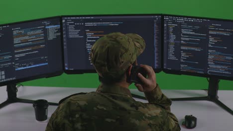 vista trasera de un hombre asiático desarrollador soldado hablando en teléfono móvil mientras escribe código con múltiples pantallas de computadora en el estudio de pantalla verde