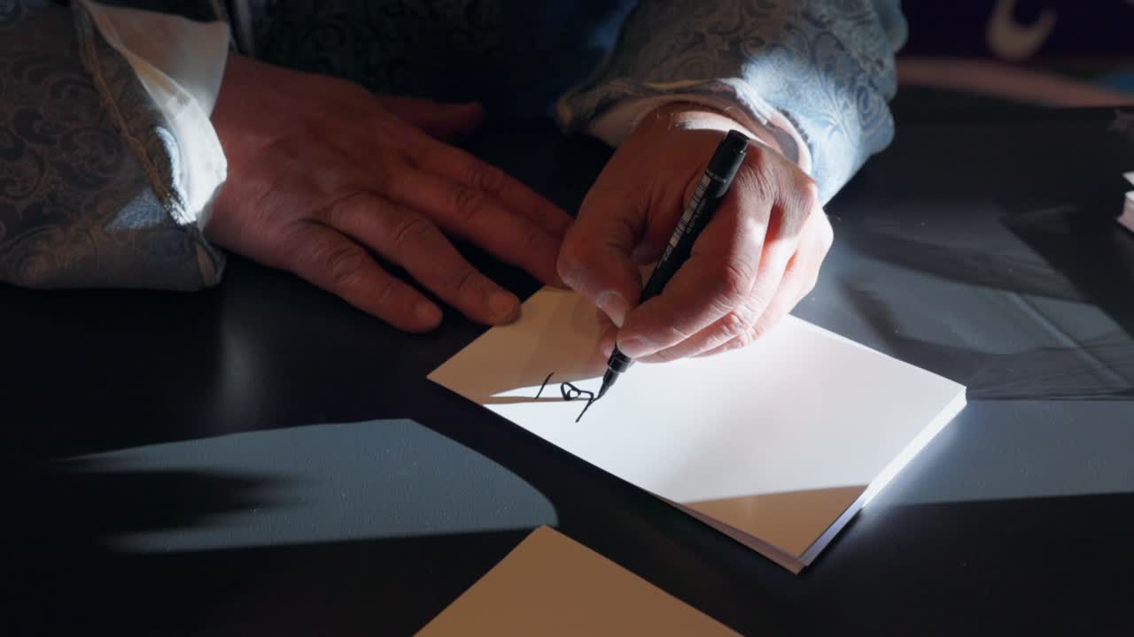 Writing Arabic calligraphy on a blank card with a black pen, wearing an ornate patterned jacket. highlighting traditional Middle Eastern art, cultural heritage, and calligraphic practice