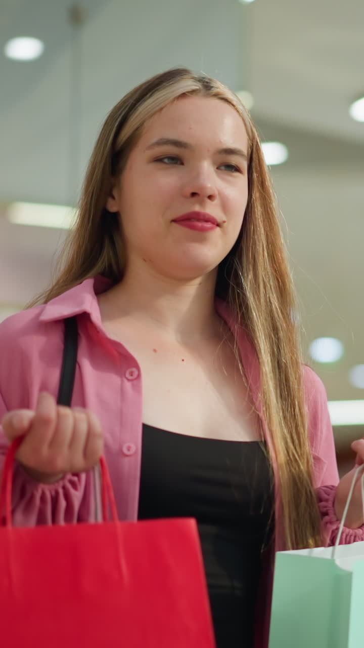 hermosa dama en vestido rosa levanta sus bolsas de compras rojas y verdes menta con una sonrisa satisfecha, tomando una respiración profunda, ella está de pie en un centro comercial bien iluminado, con otros compradores en el fondo