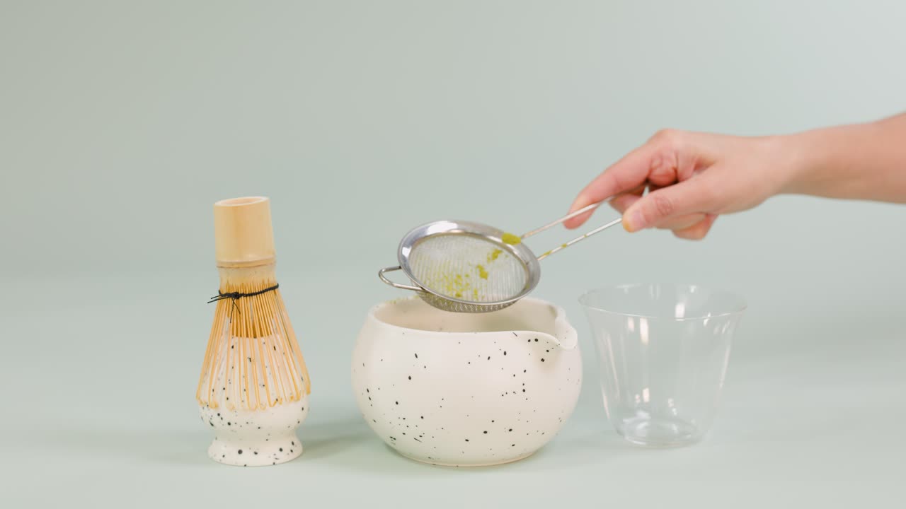 Hand sifts green matcha powder into ceramic bowl; bamboo whisk, glass cup nearby, soft lighting