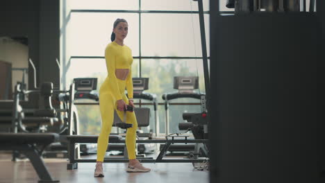 young hispanic brunette woman squats holding a dumbbell in the gym in a yellow tracksuit