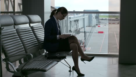 business woman in glasses, suit and heels using a tablet while sitting in the airport looking out the window while waiting for a flight