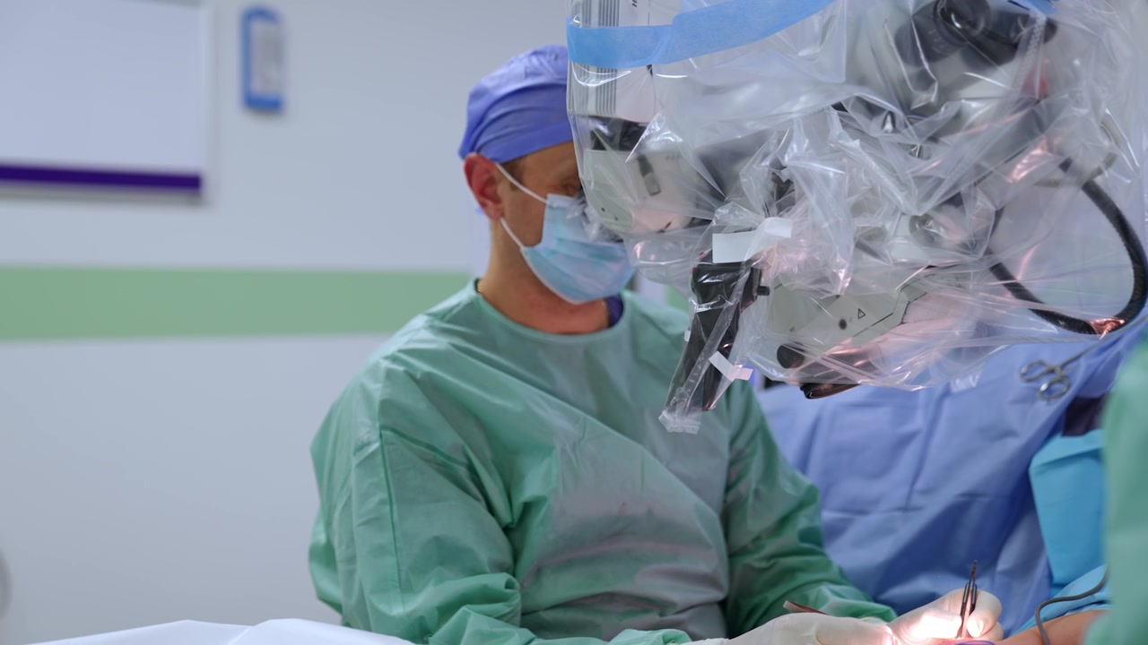 Neurocirurgião caucasiano com máscara e touca olha para o microscópio. O médico aplica instrumentos metálicos, segurando-os com as duas mãos.