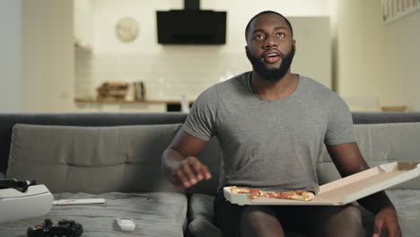 african man sitting on sofa in open kitchen. black man eating pizza.