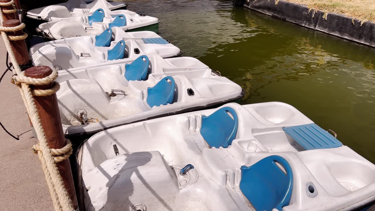 Row of white pedal boats, blue seats docked along calm green water canal, Mexico