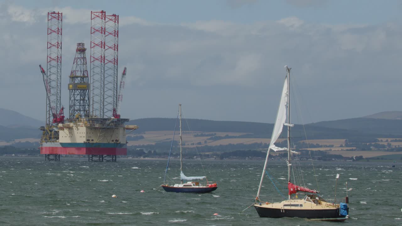 Two sailboats pass a large offshore oil rig in choppy daylight seas near Cromarty, Scotland
