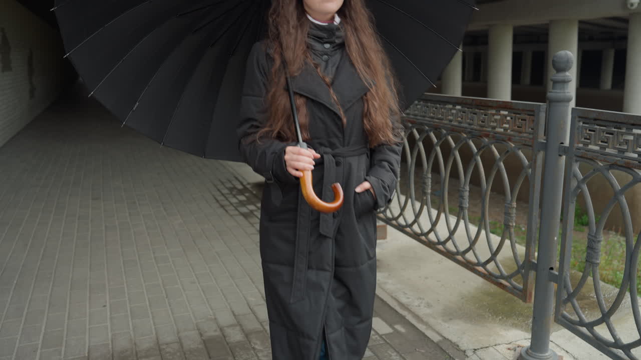 Donna che cammina per strada con un ombrello, capelli lunghi e cappotto nero, denim blu e passo disinvolto lungo una ringhiera di ferro, marciapiede bagnato e riflessi nelle pozzanghere, pioggerellina nuvolosa, tragitto pendolare spontaneo e malinconico