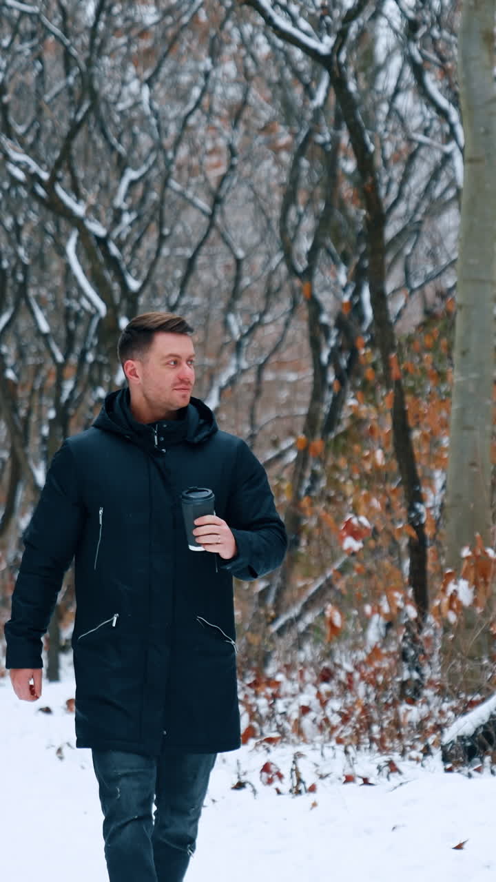 Caucasian male wearing warm winter clothes walking by the park. Man holds a paper cup in his hand and looks at his wrist watch. Vertical video