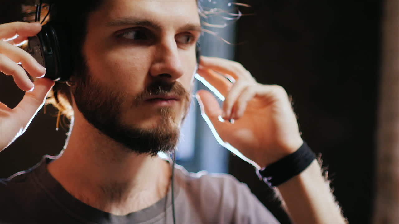 молодой хипстер с бородой слушает музыку, вокруг кирпичные стены, ветер тихонько шелестит