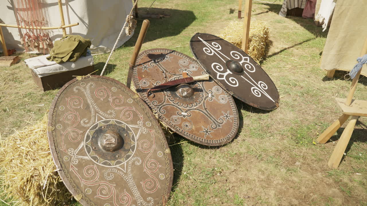 A slow dolly shot moves past a display of historical replica shields. The ornate, handcrafted shields feature intricate Celtic and Dacian patterns, resting in a reenactment camp