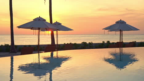 orange tropical sunset above calm sea horizon, swimming pool and beach umbrellas with mirror reflection, full frame