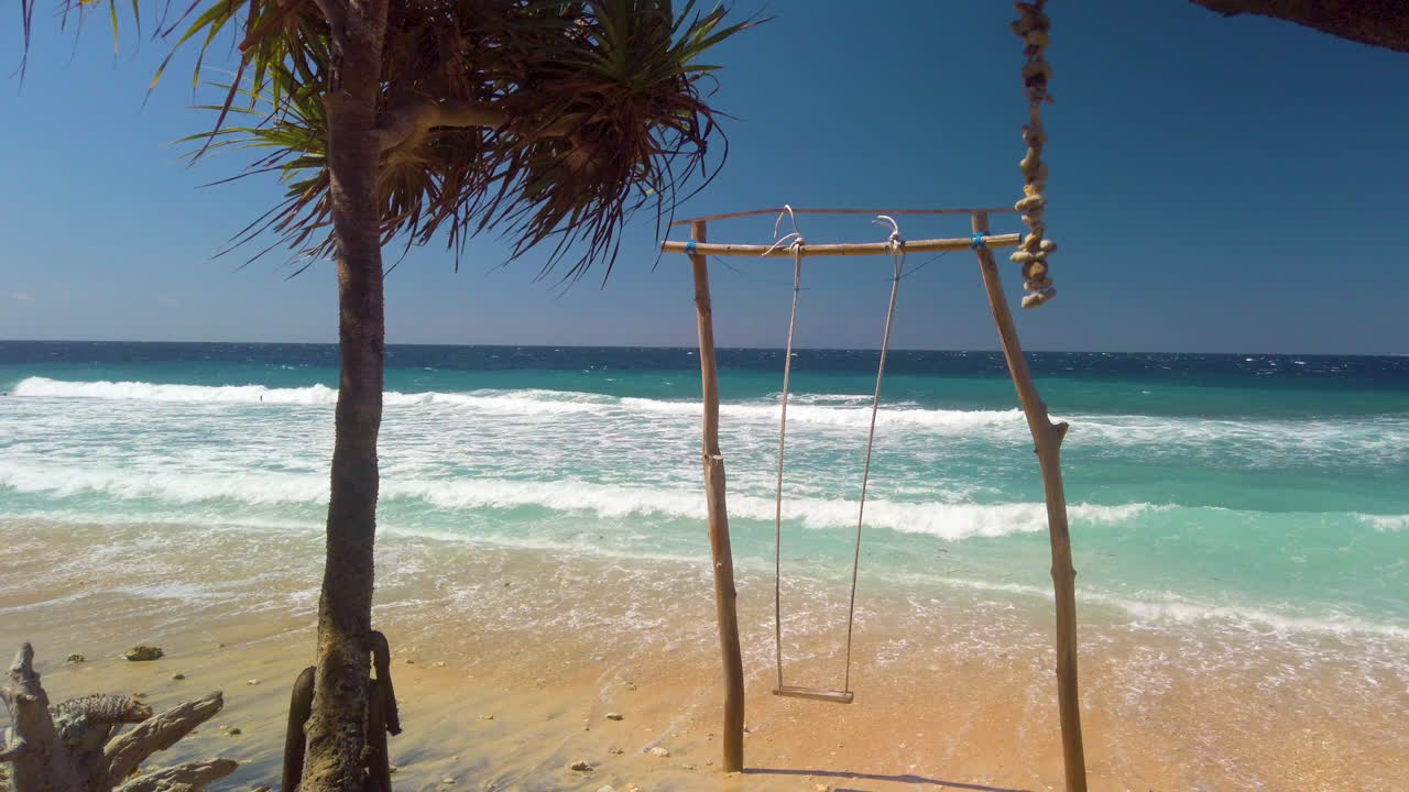 Traditional Balinese Beach Swing Hidden in Nusa Penida most tropical Island. Isolated tropical beach with palm trees and Swing with Big waves foaming in background and crushing on shore