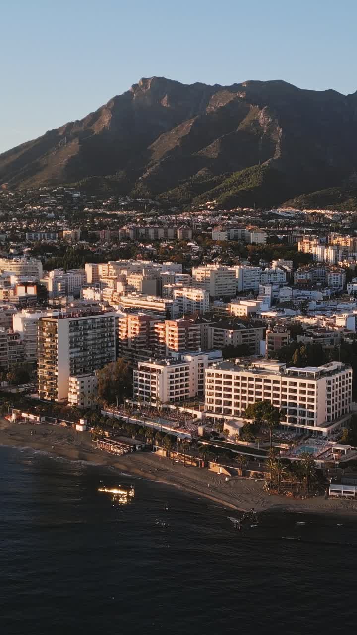 Toma aérea vertical con dron de la costa de Marbella al atardecer, con el mar Mediterráneo brillando abajo y montañas elevándose detrás, capturando el sereno lujo costero de la Costa del Sol, España.