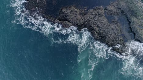 Top-down-aerial-tracking-forward-over-vibrant-blue-sea,-and-revealing-a-low-lying-rock-pool-where-sea-water-is-breaking-its-defence