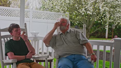 two family members are sitting on a porch, savoring ice cream treats. the elder man shares a moment with a young boy as they relax in a serene garden setting.