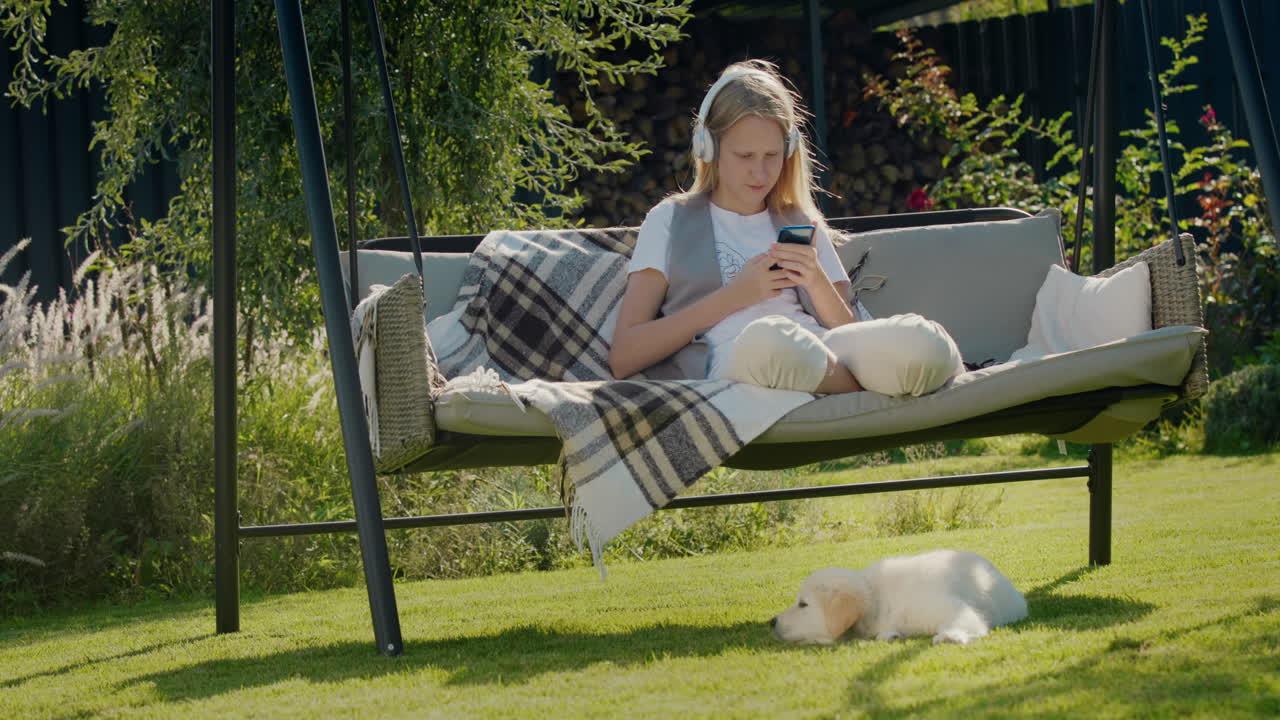 En teenagepige hviler sig på en havesofa og bruger en smartphone. Hendes hvalp sidder ved siden af hende.