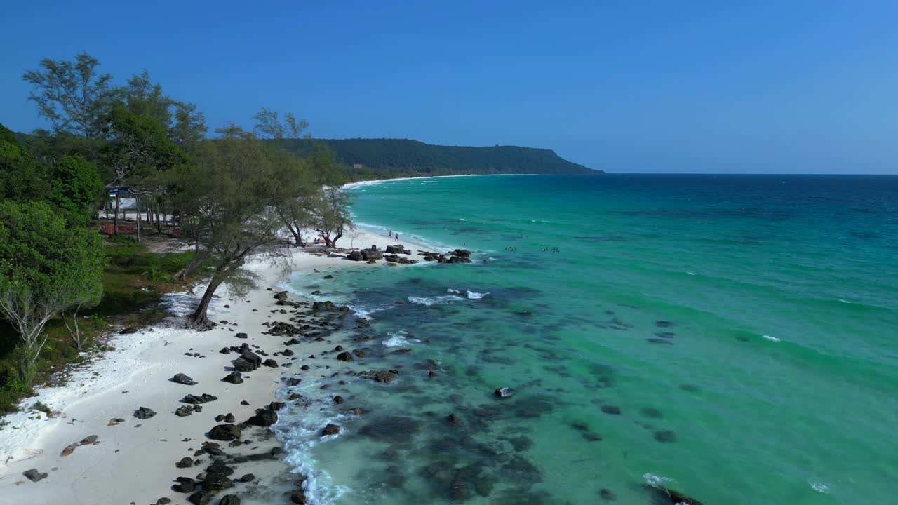 Black Stones in turquoise water gently washing the white sand beach of Koh Rong island, cambodia, on a sunny day with clear blue sky. Perfect aerial view flight static tripod hovering drone