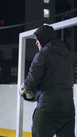 futbolista patea una pelota de fútbol durante la práctica nocturna cerca del poste de la portería en un campo urbano iluminado, el jugador demuestra enfoque bajo la iluminación del estadio con edificios residenciales en el fondo