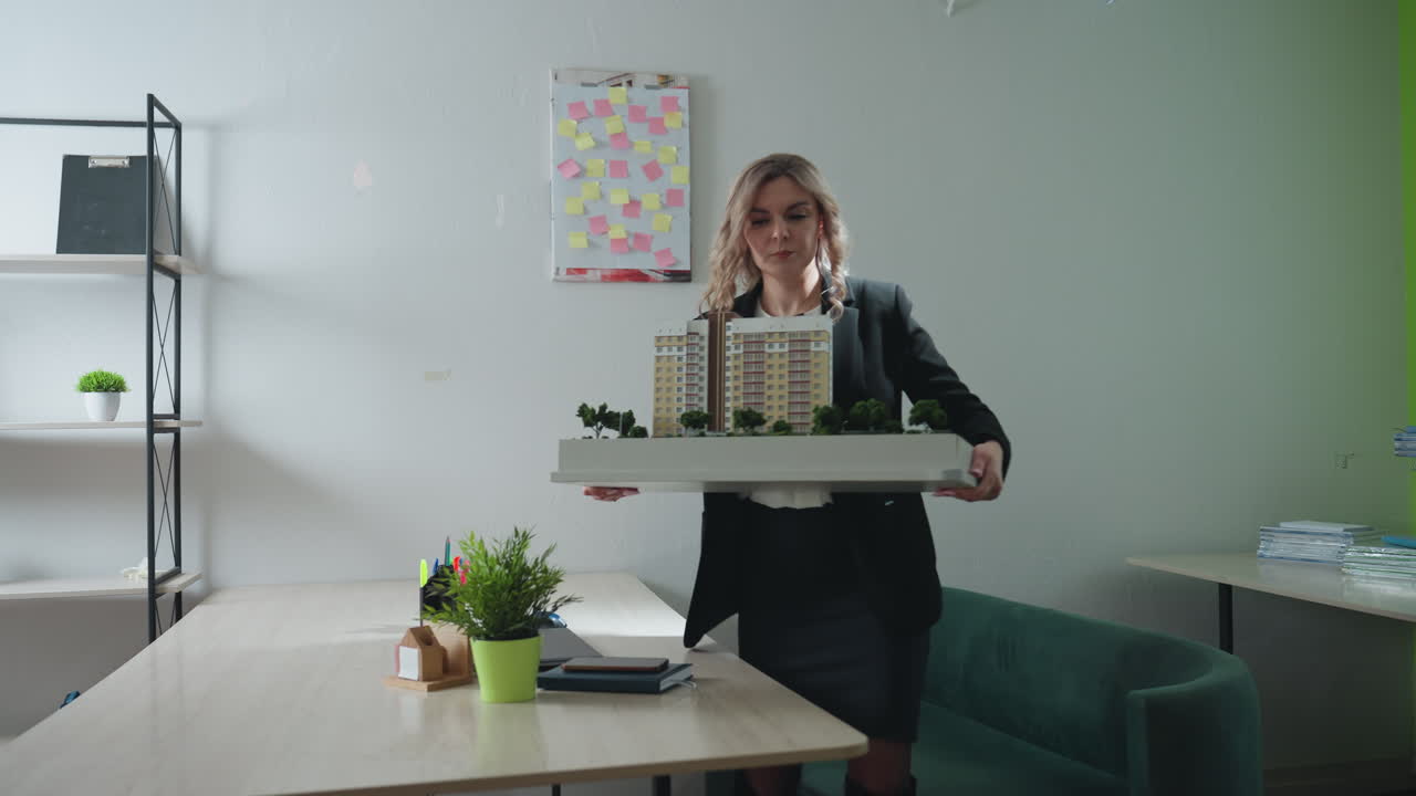 Real estate mogul lifts architectural building model from desk and exits office, modern interior features green couch, shelf with plants and files, creative board with sticky notes on white wall